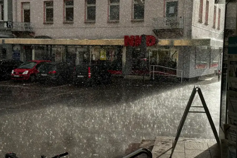 Hevige regenbui op een straat met geparkeerde auto's en een winkelpand, gezien vanuit een deuropening